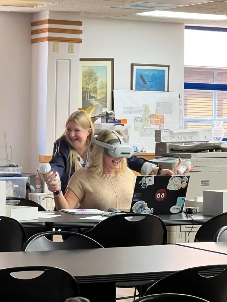A joyful woman uses a VR headset and controllers in an office, while another woman stands behind her, smiling.