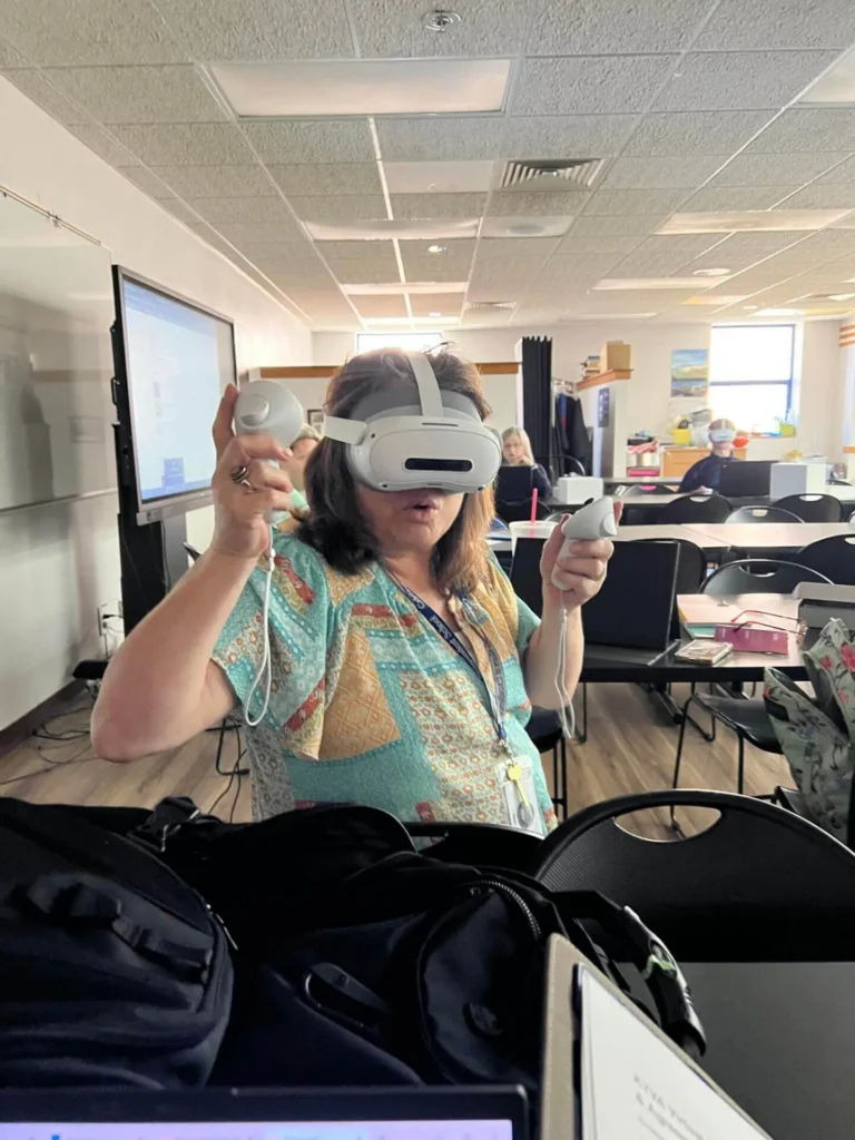 A woman in a colorful shirt uses a virtual reality headset and controllers in a bright classroom setting.