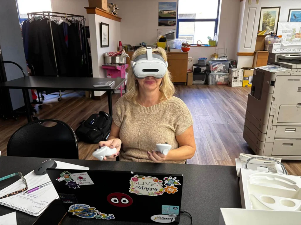 A woman sits at a cluttered desk in an office wearing a VR headset and holding controllers.