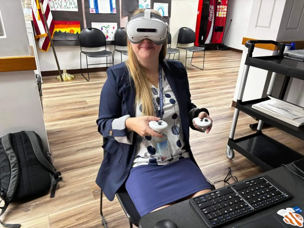A woman in a polka dot shirt and blue skirt wears a VR headset and holds controllers, smiling in an office with chairs, a backpack, and a keyboard nearby.